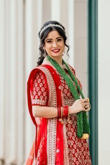 Indian bride with bridal makeup wearing red lehenga, green necklace, tiara, and bold red lipstick for traditional wedding.