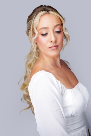 Bride with blonde hair in romantic ponytail, soft glam wedding makeup, glowing skin, long lashes, and neutral lip color.