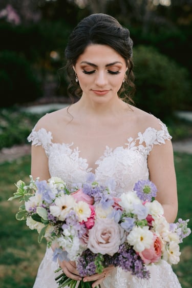 Bride with romantic bridal updo, soft wedding makeup, long lashes and neutral tones, holding pastel bouquet outdoors.