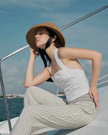 Model in white resort wear sits on a yacht in Bali, holding a hat.