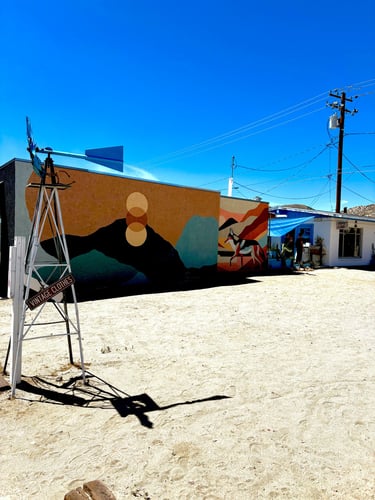 A vintage windmill stands before a desert building featuring a vibrant mountain landscape mural and blue sky.