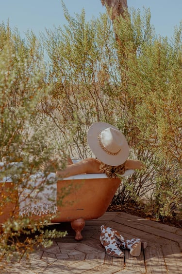 Woman in a wide-brimmed hat relaxing in an outdoor copper bathtub in the desert.