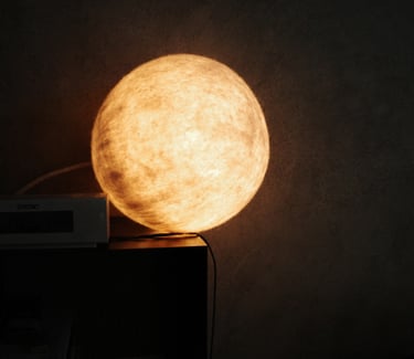 A glowing round moon texile lamp with warm yellow light on a dark nightstand.
