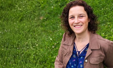 Smiling woman with curly hair wearing a brown jacket outdoors in a green grass field.