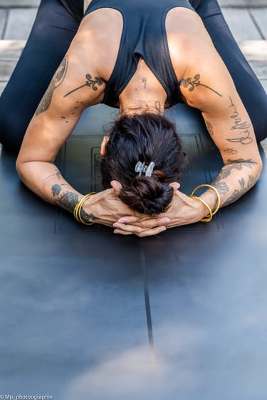 A tattooed woman practices child's pose during a yoga session on a black exercise mat.