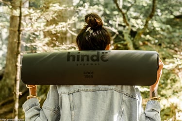 A woman in a denim jacket carrying a black Hindë yoga mat on her shoulders in a sunny forest.