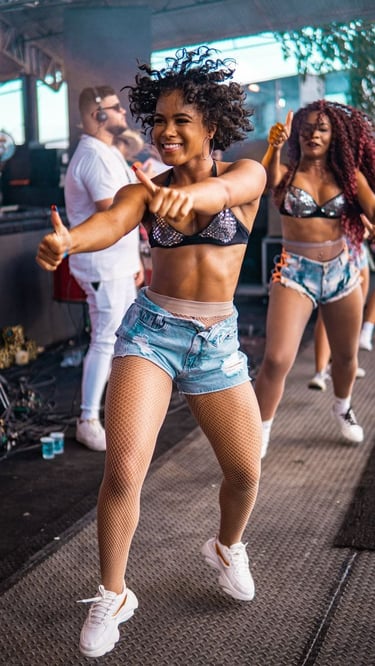 Duas mulheres negras dançam em ambiente festivo ao ar livre, vestindo top preto e short jeans.