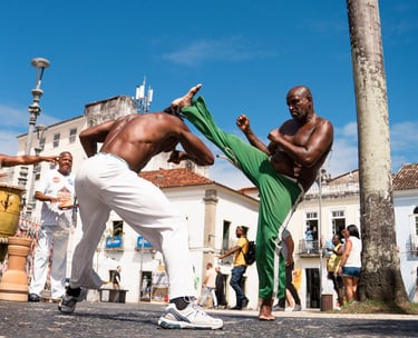 Foto de dois homens negros praticando capoeira em uma praça, com um golpe de perna alta e esquiva.