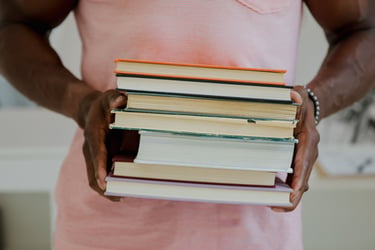 Foto de uma pessoa de pele retinta com camiseta rosa-claro segurando uma pilha de livros.