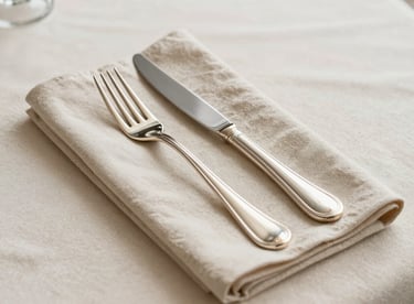 A detailed shot of a linen napkin and polished silver cutlery on a cream-colored tablecloth.