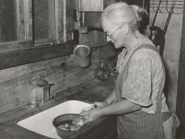 Foto vintage en blanco y negro de una mujer mayor lavando en el fregadero de una cocina rústic