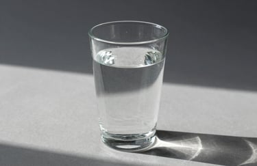 A close-up photograph of a clear glass of water on a minimalist grey surface, casting geometric shadows in a bright, airy North American studio. Symbolizing clarity and essential intent.