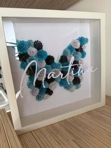 cornice bianca con all' interno farfalla realizzata con fiori di carta sui toni dell' azzurro e  Martina in bianco sul vetro
