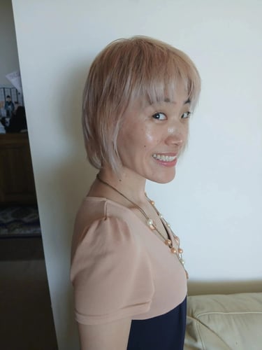 Smiling woman with a short blonde bob hairstyle wearing a peach top and pearl necklace.