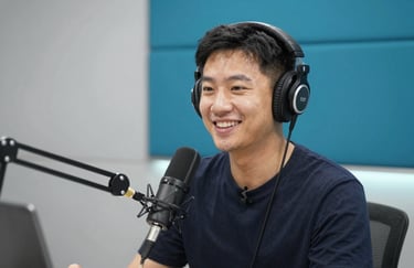 A cheerful radio presenter with headphones talking into a mic, in a clean studio with #3A7CA5 wall panels and bright lighting.