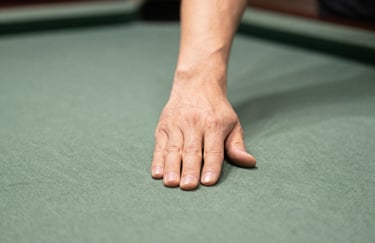 A close-up photograph of a player's hand in a professional bridge position on a billiard table. The lighting emphasizes the focus and precision, with a soft-focus background of a sage green table.