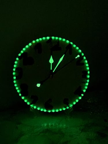 Glow in the dark wall clock with green luminescent LED lighting and glowing hands in a dark room.