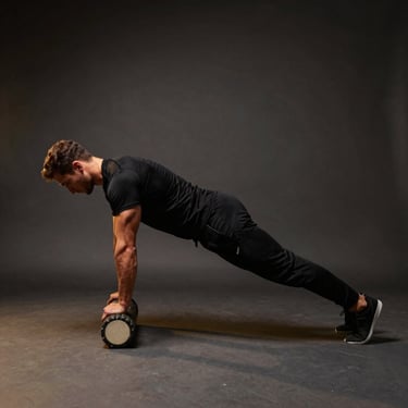 A male athlete performing mobility work with a foam roller in a dark, clean studio. He is wearing long black athletic trousers and a black shirt. Subtle gold lighting. Global / Performance Athletics.