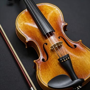 A master-crafted violin and its bow, featuring light oak wood tones and high-gloss varnish, set against a dark background.