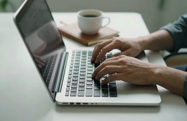 A close-up of hands typing on a modern laptop keyboard with a coffee cup and a leather-bound notebook on a clean surface, in a professional workspace with muted green accents.