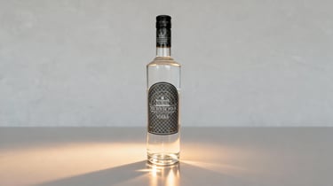 Een hoge glazen fles The House Pour Vodka op een witte ondergrond met warme lichtaccenten.