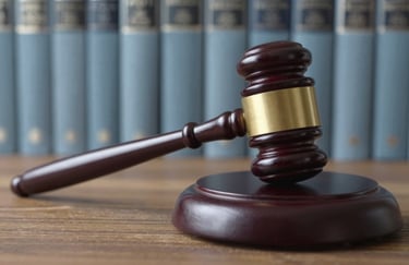 A macro photograph of a legal gavel resting on its sound block on a wooden surface. The background features bookshelves with muted steel blue spines.
