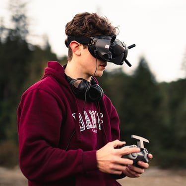 A man wearing FPV goggles and holding a remote controller to fly a drone outdoors.