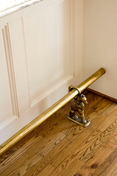 Antique brass foot rail with ornamental griffin bracket on hardwood floor in an elegant interior.