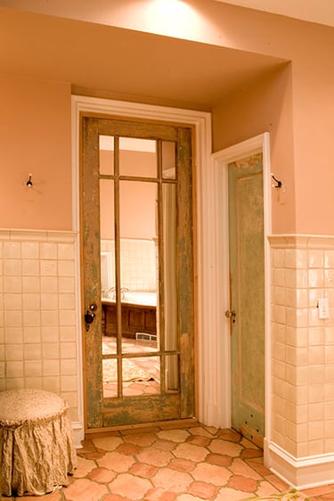 Vintage distressed wood french door with glass panes in a rustic bathroom with terra cotta tile.