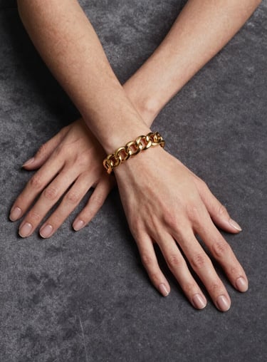 A minimalist portrait of a woman wearing a heavy gold link bracelet, her hands resting on a dark slate grey velvet surface.