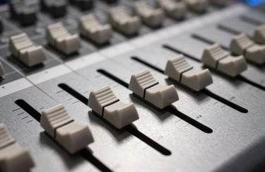 A macro shot of professional studio audio faders on a mixing desk. The focus is sharp on the metal textures with soft-white light illuminating the equipment in a European sound suite.