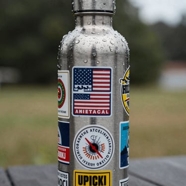 A close-up of a stainless steel water bottle covered in custom stickers, with fresh water droplets on the surface to demonstrate waterproof qualities, North American outdoor setting.