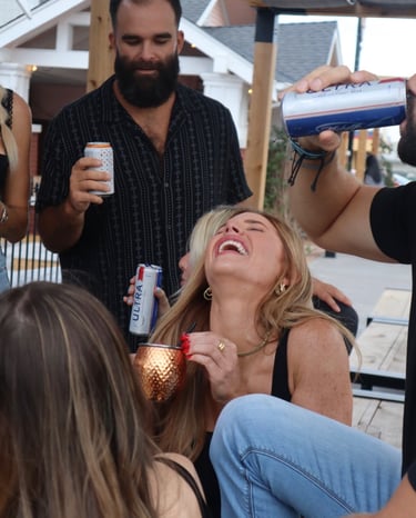 A group of friends laughing and drinking beer and cocktails at an outdoor patio bar.