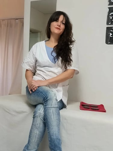 Tirzha masajista profesional en Flores, sentada en la camilla luciendo ambo blanco y jeans en su gabinete.