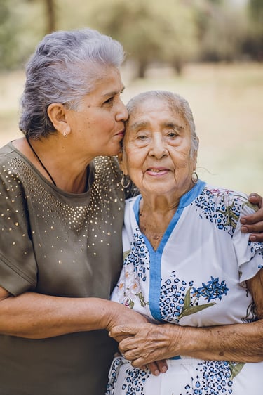 Una mujer mayor besando la frente de su madre anciana al aire libre, mostrando amor y cuidado