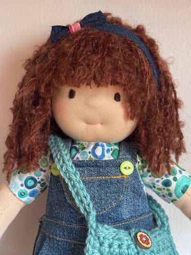 Muñeca Waldorf hecha a mano con cabello castaño rizado, overol de mezclilla y una bolsa azul tejida