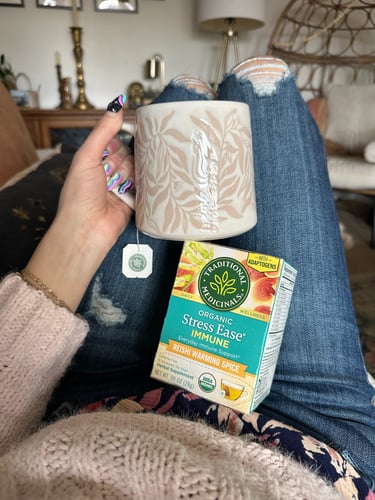 photo of someone's hand holding a mug in front of their knees laying down, a box of Traditional Medicinals tea underneath