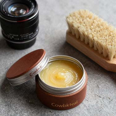 A close-up photograph of premium leather wax jars and a soft horsehair brush on a grey stone surface, professional product photography for a Swedish cobbler service.