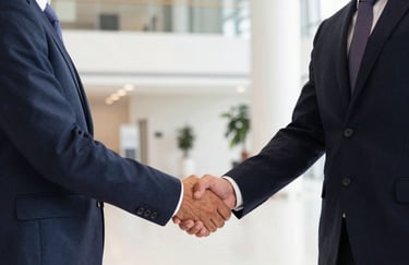 Two professionals in elegant business attire shaking hands in a brightly lit, modern lobby in Valencia. Iberian / Spanish professional context. Lighting includes subtle fuchsia hues.