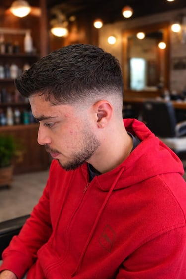 Young man with a fresh fade haircut and beard, wearing a red hoodie in a barbershop, showcasing modern men's hairstyle.