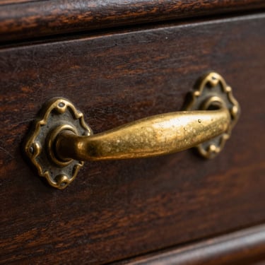 Macro photography of an ornate antique brass drawer handle, showing fine details and a gentle aged patina on dark wood.