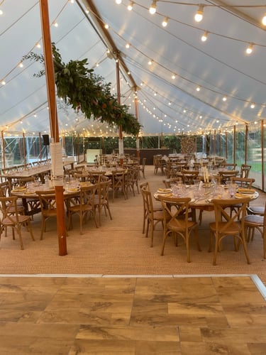 Elegant outdoor wedding reception tent with bistro lighting, wooden cross back chairs, and floral greenery.