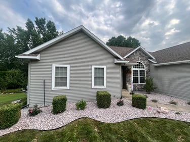A ranch style house with grey vinyl siding, stone accents, and pink gravel landscaping.