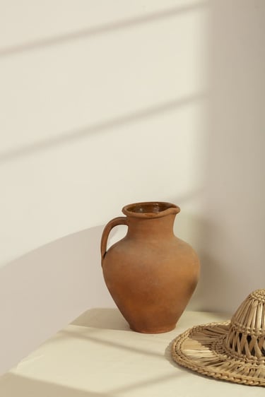 Jarra rústica de barro terracota sobre una mesa minimalista, acompañada de un sombrero de paja.