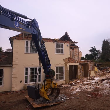 Blue excavator arm demolishing a two-story residential house with debris and rubble on the construction site.