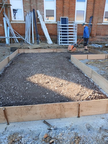 A construction worker uses a plate compactor to level gravel within wooden formwork for a concrete foundation.