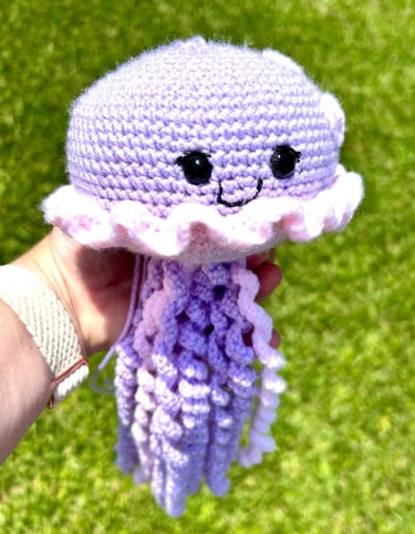 Handmade purple and pink crochet jellyfish amigurumi toy with curly tentacles held outdoors over green grass.