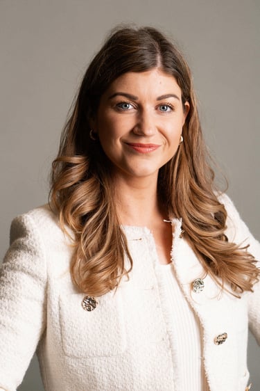 Professional headshot of a smiling woman with wavy brown hair wearing a white tweed blazer.