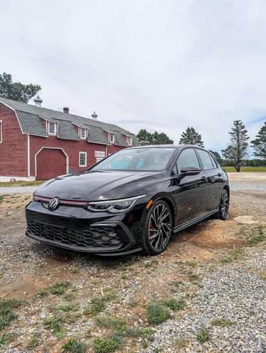 A black Volkswagen Golf GTI parked on a gravel path in front of a classic red farm barn.