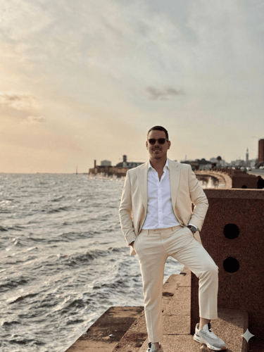 Carlos Assies in a cream-colored linen suit and sunglasses poses on a coastal boardwalk at sunset.
